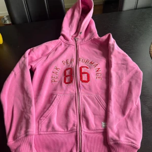 Rosa hoodie från Peak Performance - Säljer en rosa hoodie från Peak Performance med dragkedja och huva. Tröjan har broderad text och siffran 86 framtill, samt två stora fickor. Mjuk insida och klassisk passform, perfekt för chill dagar.