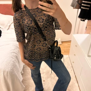 Blommig spetsblus - Säljer en elegant blommig spetsblus i mörkblått och beige. Blusen har korta ärmar och en fin genombruten design som ger en stilren look. Perfekt för att lyfta vilken outfit som helst.