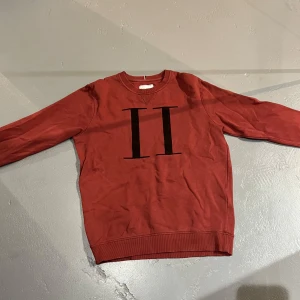 Röd sweatshirt från Les Deux - Säljer en snygg röd sweatshirt från Les Deux med ett stilrent svart tryck på framsidan. Tröjan har långa ärmar och en rund halsringning. Perfekt för en avslappnad stil.