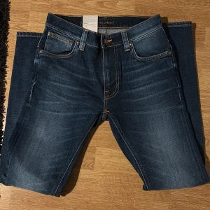 Blå jeans från Nudie Jeans - Snygga blå jeans från Nudie Jeans i 100% ekologisk bomull. De har en klassisk femficksdesign med orange sömmar och en läderpatch med logotyp baktill. Perfekta för en stilren look.