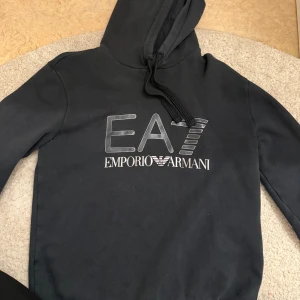 Svart hoodie från Emporio Armani - Snygg svart hoodie från Emporio Armani med EA7-logga på bröstet. Den har en klassisk huva med dragsko och långa ärmar. Perfekt för en stilren och bekväm look.