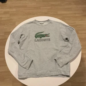 Grå långärmad tröja från Lacoste - Säljer en grå långärmad tröja från Lacoste med det ikoniska gröna krokodilmärket på bröstet. Tröjan är i en mjuk bomullsmix och har en avslappnad passform. Perfekt för en casual look.