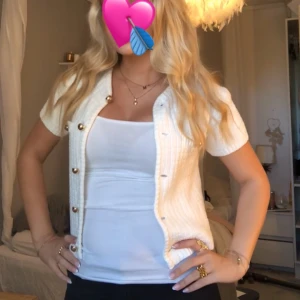 Vit kortärmad kofta från H&M - Säljer en stilren vit kofta från H&M med korta ärmar och dekorativa guldknappar. Koftan har två små fickor framtill och en texturerad yta som ger en elegant touch. Perfekt för att lyfta din outfit! 🤍
