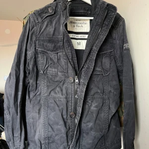 Field jacket från Abercombie and fitch - Säljer en svart vintage jacka från Abercrombie & Fitch med dragkedja och knappar. Jackan har flera fickor och en broderad logga på ärmen. 