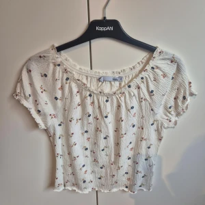 Vit blommig topp från Lager 157 - Vit topp med blommigt mönster i rött och blått. Toppen har korta ärmar och en elastisk halsringning som ger en bekväm passform. Perfekt för en avslappnad stil.