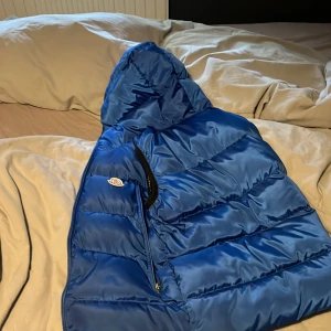 Blå pufferjacka från Moncler - Säljer en snygg blå pufferjacka från Moncler med svart foder och logotypdetaljer. Jackan har en huva och dragkedja framtill. Perfekt för kyliga dagar när du vill hålla stilen.