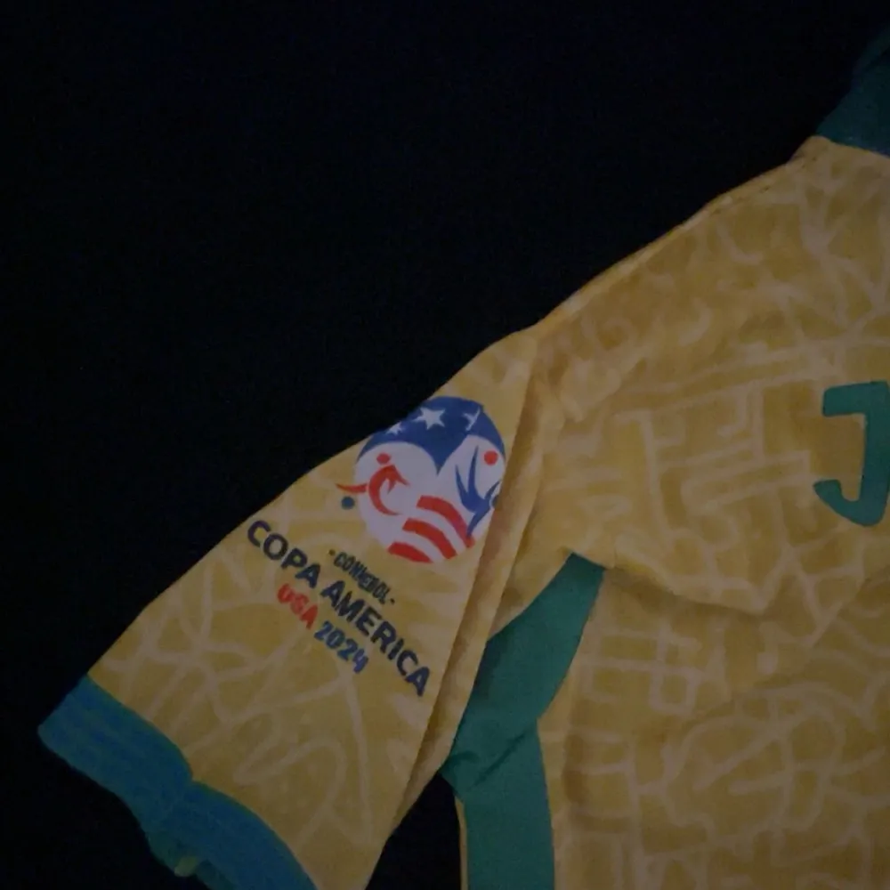 Säljer en gul Brasilien fotbollströja från Nike med gröna detaljer och nummer 7. Tröjan har korta ärmar och ett tryck för Copa America USA 2024 på ärmen. Perfekt för fotbollsfans! Tröjan är liten i storleken, kom privat för mer information, helt ny. T-paidat.