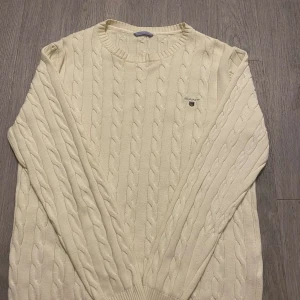 Vit stickad tröja från GANT - Säljer en stilren vit stickad tröja från GANT med kabelstickat mönster. Tröjan har långa ärmar och en rund halsringning. Perfekt för en klassisk och tidlös look. GANT-logotypen är broderad på bröstet.