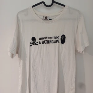 A bathing ape X mastermind tee - Säljer en vit t-shirt från Aape med tryck av 'mastermind A BATHING APE' och en dödskalle. T-shirten har en rund halsringning och korta ärmar. Perfekt för en avslappnad stil.