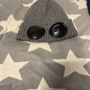 Grå ribbad mössa från C.P. Company - Säljer en grå ribbad mössa från C.P. Company med unika glasögondetaljer framtill. Perfekt för att hålla stilen under kyliga dagar. Mössan är i enhetlig storlek och har en modern design. Och är äkta det finns Scan kod och sånt. 