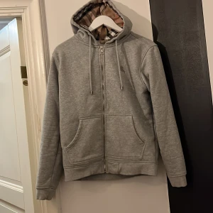 Grå hoodie från Burberry - Säljer en stilren grå hoodie från Burberry med klassiskt rutigt mönster i fodret. Tröjan har dragkedja framtill och en bekväm huva. Helt äkta köpt från Burberry outlet och säljer därför billigt. Nästan nyskick och storlek M. Sitter lite litet så kan även passa storlek S. Kontakta för frågor eller fler bilder 😁Säljer pga för att den var för liten på mig.