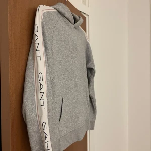 Grå hoodie från GANT - Säljer en stilren grå hoodie från GANT med vita och rosa detaljer längs ärmarna. Tröjan är endast använd 2-3 gånger. Tröjan kostar ca 1000 kr nypris. Pris går att diskutera