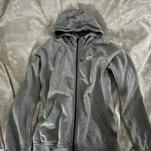 Grå hoodie från Nike - Säljer en grå hoodie från Nike med dragkedja och huva. Perfekt för en avslappnad stil. Den har långa ärmar och en klassisk passform. Nike-loggan är tryckt på bröstet. Skön och stilren design för alla tillfällen.