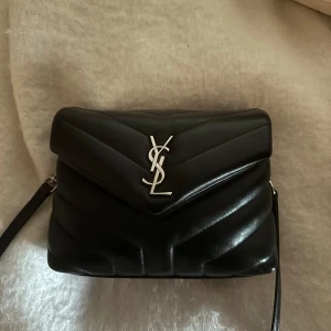 Yves Saint Laurent loulou väska - Snygg svart axelväska från Saint Laurent i quiltat skinn. Väskan har en elegant design med silverfärgad logotyp på framsidan och en praktisk dragkedja inuti. Perfekt för att ge din outfit en lyxig touch.