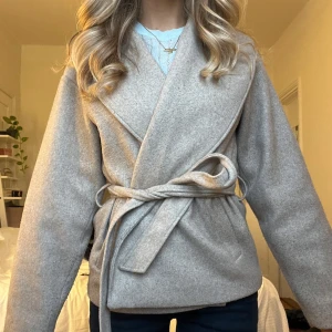 Beige omlottkappa med bälte - Snygg beige omlottjacka med bälte i midjan. Perfekt för höst och vår, med en stilren design och långa ärmar. Jackan har en elegant passform och är tillverkad i ett mjukt material som ger en bekväm känsla.