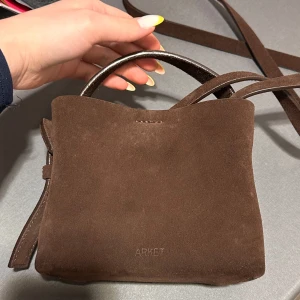 Brun mocka handväska - Snygg brun handväska i mocka med ett stilrent och minimalistiskt utseende. Väskan har ett handtag och en dekorativ knäppning på sidan. Perfekt för att bära med sig det viktigaste med stil. Väskan har en liten mörk fläck på baksidan och det är vanligt att sånna dyker upp på mocka väskor då mocka ändras med tiden samt knappt tål nånting som tillexempel vatten