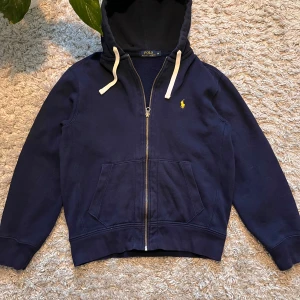 Mörkblå hoodie från Polo Ralph Lauren - Säljer en snygg mörkblå hoodie från Polo Ralph Lauren med dragkedja och klassisk gul logga på bröstet. Den har en justerbar huva med vita snören och praktiska fickor framtill. Perfekt för en avslappnad stil.  , inga fläckar, dock liten slitning på ena handen, skickar bild på de privat om du är intresserad. Annars väldigt skön och snygg