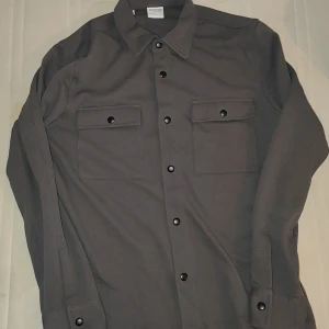 Grå overshirt från Selected Homme - Snygg grå overshirt från Selected Homme med långa ärmar och stilrena svarta knappar. Två praktiska bröstfickor med lock framtill. Perfekt för lager-på-lager-stil. Väldigt snygg till sommaren och helt oanvänd nästan så den har inga tecken på användning direkt sådär.