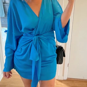 Blå omlottklänning med knyt - Snygg blå omlottklänning med knyt i midjan. Klänningen har långa ärmar och en v-ringad design som ger en elegant look. Perfekt för att sticka ut med sin klara färg och stilrena detaljer.