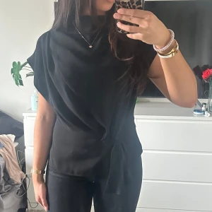 Svart topp med knyt - Snygg svart topp med knyt i midjan och korta ärmar. Perfekt för en stilren look. Toppen har en elegant design som passar till många tillfällen.