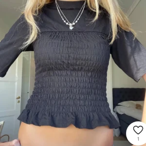 Svart smockad topp - Säljer en svart smockad topp med korta ärmar och volangdetalj vid nederkanten. Perfekt för en stilren look. Supernice!!!!💞💞💞