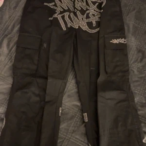 Svarta cargopants med graffiti-design - Snygga svarta cargopants med unik graffiti-design i vitt. Byxorna har flera fickor och en loose passform, perfekt för en avslappnad stil. Detaljerna ger en cool och urban känsla.