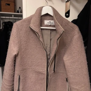 Beige jacka John henric - Snygg beige jacka från John henric, i ull material på utsidan. Dragkedja framtill och två praktiska fickor. Jackan har en stilren design med långärmade ärmar och en mjuk, texturerad yta. Perfekt nu för våren.