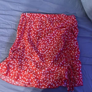 Röd blommig kjol med volang - Säljer en söt röd kjol med vita blommor och volangdetaljer. Kjolen har en dragkedja på sidan och en kort längd som passar perfekt för varmare dagar. Den är gjord i ett lätt och luftigt material. Inte använd speciellt mycket! 