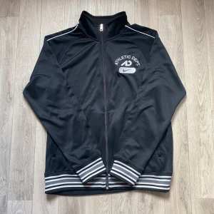 Nike zip - Säljer en svart träningsjacka från Nike med dragkedja och Athletic Dept-tryck på bröstet. Jackan har ribbade muddar och krage samt vita detaljer på ärmarna. Perfekt för sportiga aktiviteter eller en avslappnad stil. Priset kan diskuteras 