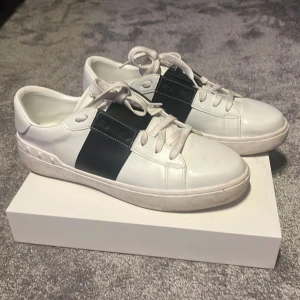 Valentino open - Snygga vita sneakers från Valentino med en bred svart rem över ovansidan. Skorna har snörning och en unik detalj med nitar runt hälen. Perfekta för en stilren look.