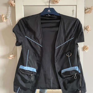 Svart  väst- jacka från PrettyLittleThing - Säljer en svart kortärmad jacka från PrettyLittleThingw med ljusblå detaljer. Jackan har dragkedjor på fickorna och en cool design med ringdetaljer. Perfekt för en trendig look! Helt ny oanvänd.storlek xs.kan möta er i Stockholm T-Centralen utanför Åhléns 