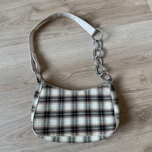 Rutig axelväska med kedjedetalj - Snygg rutig axelväska med en cool kedjedetalj på axelremmen. Väskan har en dragkedja upptill och ett stilrent rutigt mönster i svart, beige och brunt. Perfekt för att ge din outfit en trendig touch.