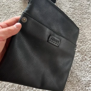 Svart axelväska från Armani (liten) i skinn - Snygg svart axelväska från Armani i syntetmaterial. Väskan har en justerbar axelrem och en praktisk dragkedja upptill. Perfekt för att bära med sig det nödvändigaste med stil.