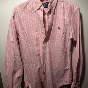 Rosa vit randig Ralph Lauren skjorta - Säljer nu min Ralph Lauren skjorta som är vit och rosa randig med en brun logga. Bra skick, storlek M och riktig fin till sommaren!
