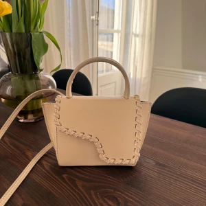 Beige handväska med flätad detalj - Snygg beige handväska med unik flätad detalj längs kanterna. Den har ett praktiskt handtag och en avtagbar axelrem för flexibilitet. Perfekt för att ge din outfit en stilren touch.