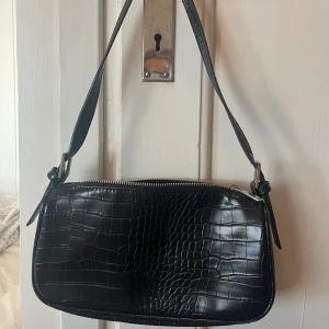 Svart axelväska med krokodilmönster - Snygg svart axelväska med krokodilmönster, dragkedja och detaljer i silver, från Gina Tricot. Perfekt för att ge en stilren touch till din outfit. Väskan har en justerbar axelrem och är rymlig nog för dina nödvändigheter.