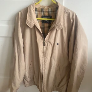 Beige jacka från Burberry - Säljer en klassisk beige jacka från Burberry med ikoniskt rutigt foder. Jackan har dragkedja framtill och en broderad logga på bröstet. Perfekt för en stilren look.