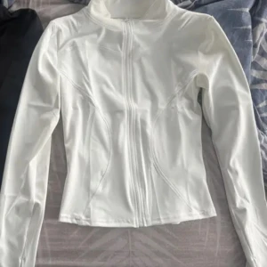 Vit långärmad jacka med dragkedja - Snygg vit jacka med lång ärm och dragkedja framtill. Perfekt för en stilren look. Jackan har en figurnära passform och är tillverkad i ett mjukt material.