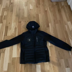 Svart jacka från Sail Racing, stl 160 - Säljer en svart jacka från Sail Racing med huva och dragkedja med en logotyp på bröstet.