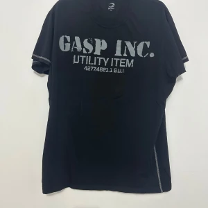 Svart t-shirt från GASP - Säljer en svart t-shirt från GASP med tryck 'GASP INC. UTILITY ITEM' på framsidan. T-shirten har korta ärmar och rund halsringning. Perfekt för en avslappnad stil.