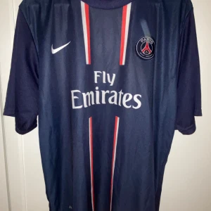 Blå PSG fotbollströja med Ibrahimović 18 - Säljer en blå PSG fotbollströja från Nike med korta ärmar. Tröjan har 'Fly Emirates' tryckt på framsidan och 'Ibrahimović 18' på baksidan. Den har röda och vita detaljer och är tillverkad i Dri-FIT material för optimal komfort.