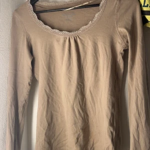 Beige långärmad topp med spetsdetaljer - Säljer en beige långärmad topp med fina spetsdetaljer runt halsringningen. Perfekt för en stilren och bekväm look. Passar bra till både jeans och kjol.