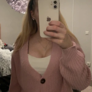 Rosa stickad kofta från Vero Moda - Säljer en söt rosa stickad kofta från Vero Moda. Koftan har en v-ringad design med stora knappar framtill. Perfekt för att hålla sig varm och stilren under kyligare dagar. Passar bra till jeans eller kjol för en avslappnad look.