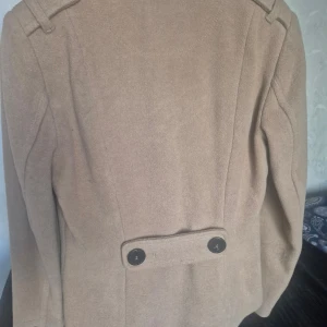 Beige kappa med ankardetalj - Snygg beige kappa med dubbelknäppning och ankardetalj på insidan. Tillverkad av 55% ull och 45% viskos, med foder i 100% polyester. Perfekt för kyliga dagar med stil. 🧥