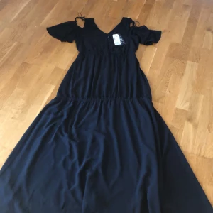 Svart långklänning från Tom Tailor - Elegant svart långklänning från Tom Tailor med v-ringning och knappar framtill. Klänningen har korta ärmar med knytning och en smickrande passform. Perfekt för en stilren look.