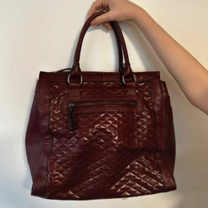 Vinröd handväska med quiltad design - Snygg vinröd handväska med quiltad design på framsidan och dragkedja. Tillverkad i syntetmaterial med två handtag för enkel bärning. Perfekt för att ge en touch av stil till din outfit.