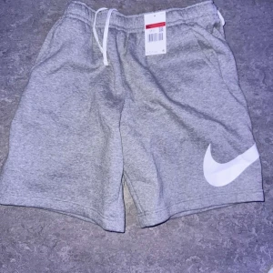 Grå shorts från Nike med vit logga - Grå mjuka shorts från Nike med vit snörning i midjan och stor vit Swoosh-logga på ena benet. Shortsen har elastisk midja och en bakficka. Perfekta för en avslappnad stil. Etiketten finns kvar.