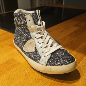 Glittriga höga sneakers med silverdetaljer - Säljer ett par höga sneakers från Philippe Model med svart och silverglittrigt tyg, vita snören och silverfärgade detaljer. Skorna har en vit sula och en rund tå. Perfekta för dig som vill sticka ut med lite extra glitter!