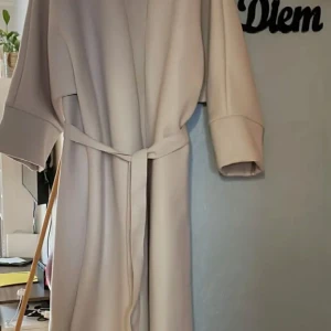 Beige kappa med bälte - Säljer en stilren beige abaya kappa med bälte i midjan och vida ärmar. Jackan har en rak och lång passform, perfekt för dig som gillar minimalistisk stil. Enkel att matcha med olika outfits. ONESZIE