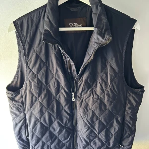 Svart quiltad väst från Oscar Jacobson - Snygg svart quiltad väst från Oscar Jacobson med dragkedja framtill. Västen har en klassisk passform och är perfekt för lager-på-lager. Tidlös design med diskret mönster och hög krage. Den är i storlek 50 vilket motsvarar en M, nypris cirka 2500